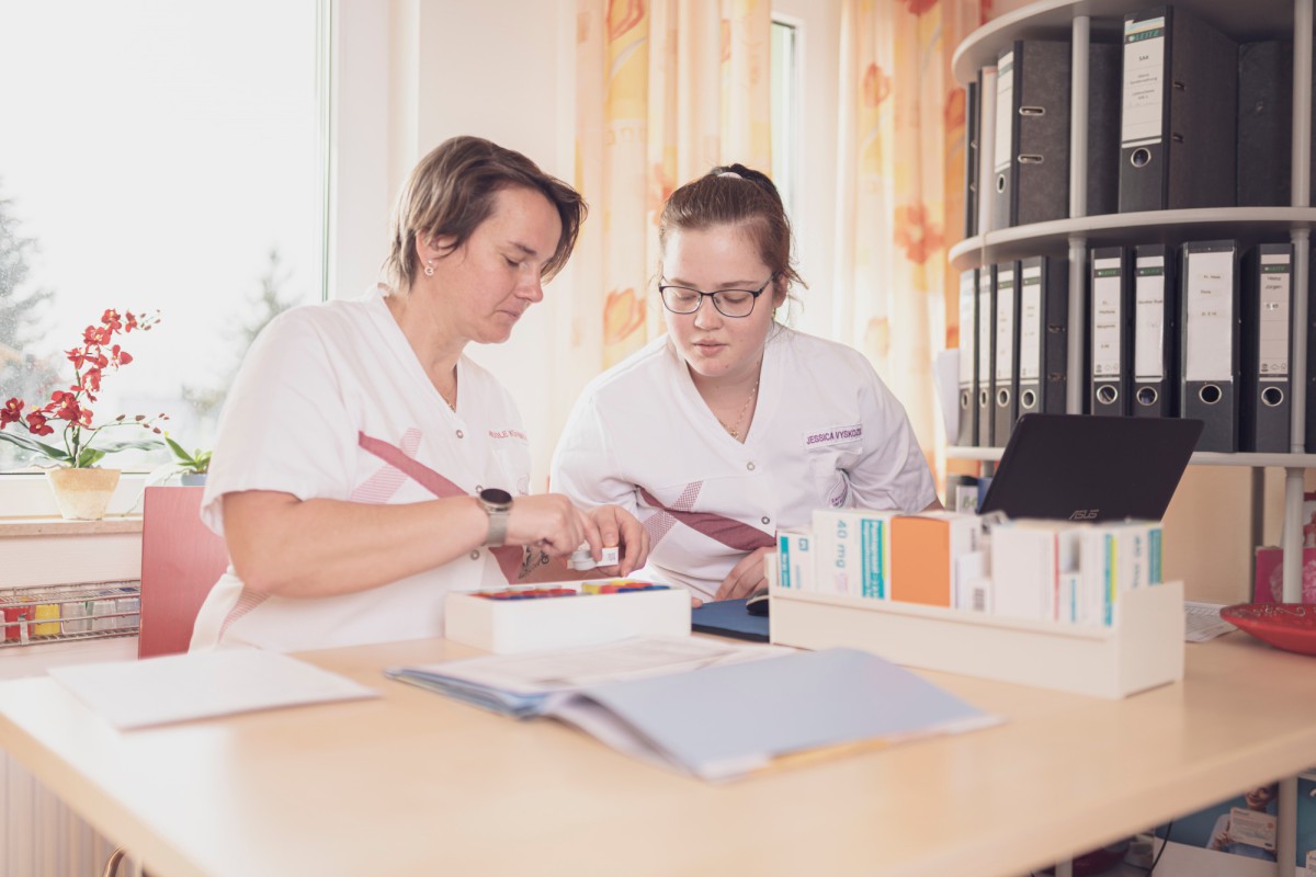 Zwei Pflegekräfte arbeiten gemeinsam an einem Tisch mit Akten, Dokumenten und Büromaterialien. Sie blicken konzentriert auf ein Dokument, während eine Person mit einer Brille etwas mit einem Stift markiert. Im Hintergrund sind Aktenstehordner und eine Topfpflanze zu sehen.