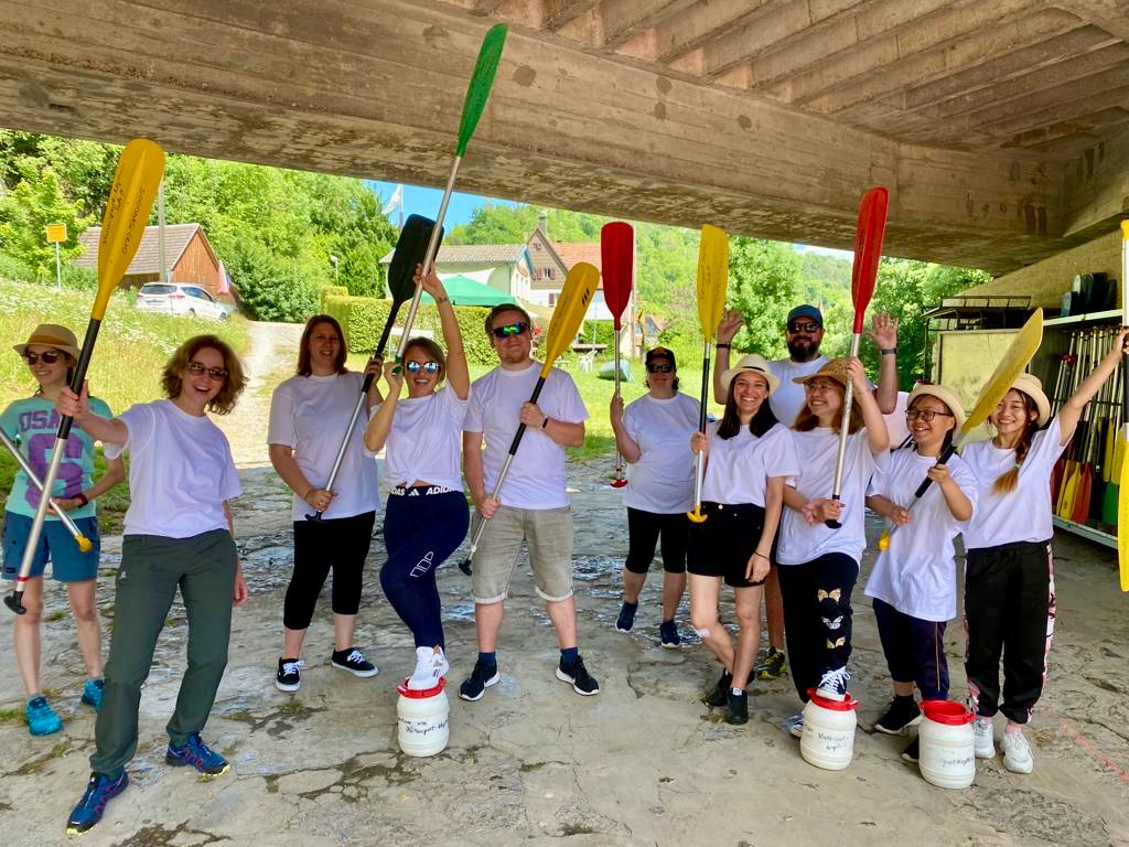 Gruppenfoto von elf Personen in weißen T-Shirts, die Paddel in verschiedenen Farben halten und vor einem hölzernen Unterstand stehen, offensichtlich bereit für eine Kanu- oder Kajakaktivität.