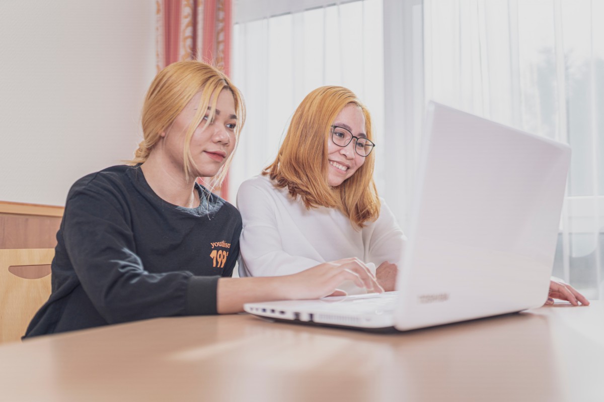 Zwei junge Personen sitzen an einem Tisch und arbeiten gemeinsam an einem weißen Laptop. Sie haben blonde Haare und lächeln, während sie konzentriert auf den Bildschirm schauen.