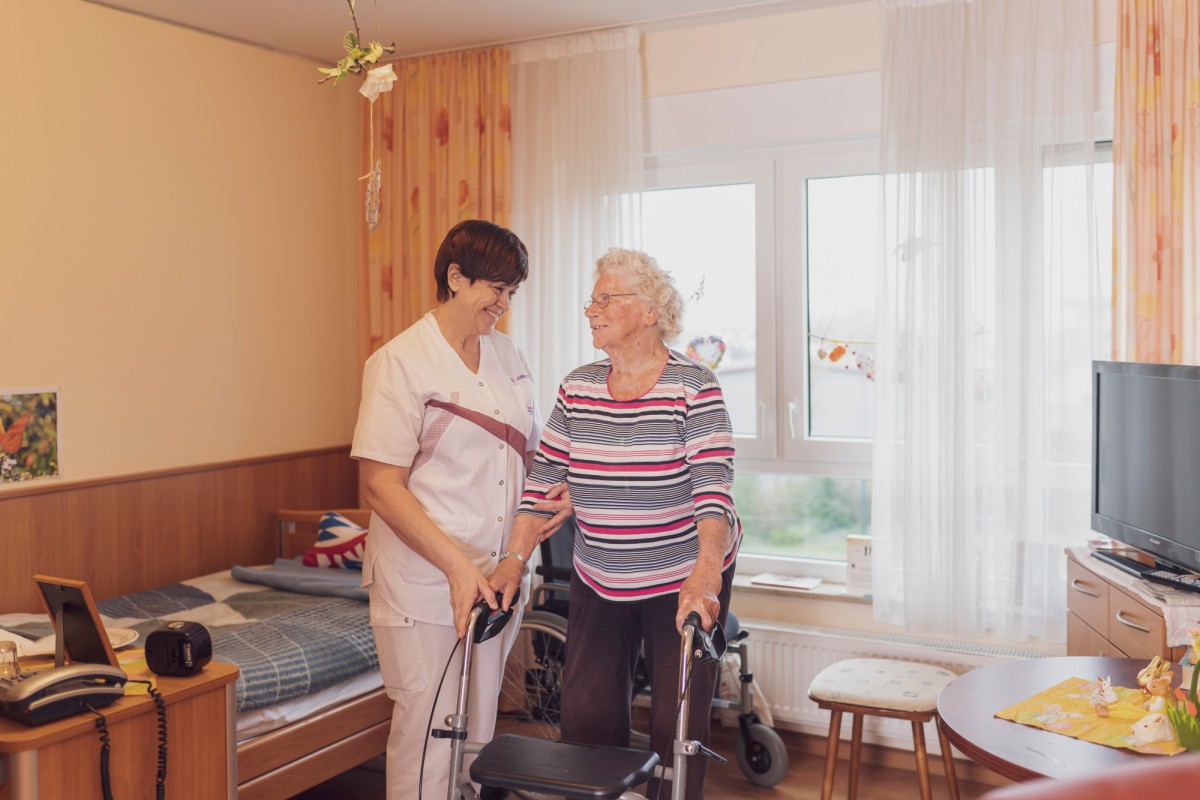 A caregiver assists an elderly woman with a walker in a bright, cozy room with wood paneling and curtains.