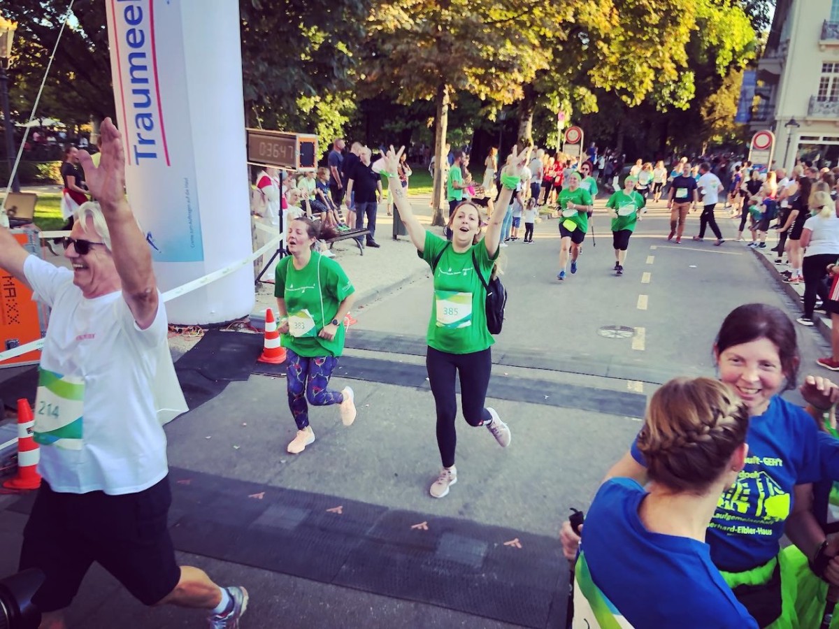 Runners in green shirts participate in a race, cheering and running under street trees and start signs.