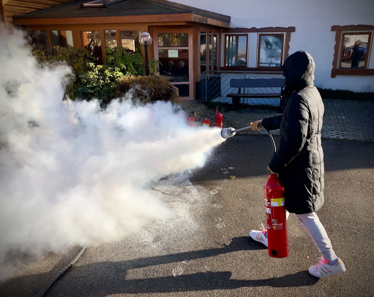 A person in a dark winter jacket and hood operates a red fire extinguisher, releasing a large white smoke plume in front of a building with wooden overhang and glass windows.