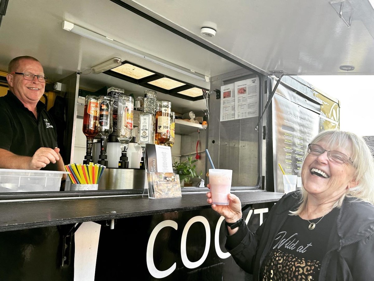 Two smiling people at a beverage stand. A man in a black shirt stands behind the counter with drink bottles, while a woman with glasses and a black top laughs while holding a drink.