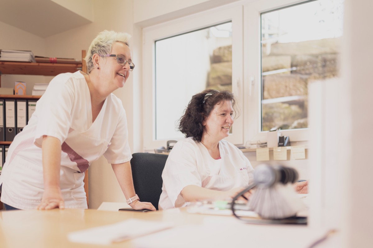 Two female employees in white professional attire work focused at a bright workspace with file folders and documents. They appear friendly and concentrated while working together.