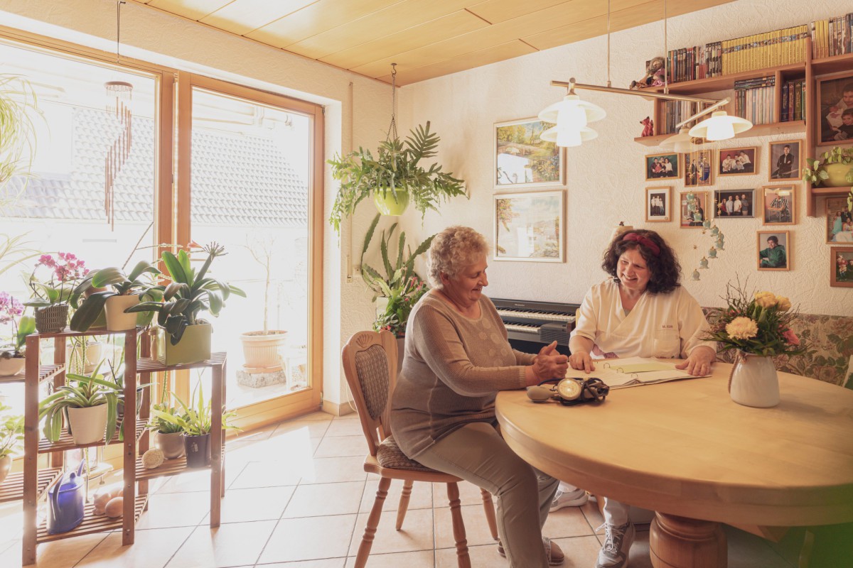 Two women sitting at a light wooden table in a sunlit room with plants and bookshelves.