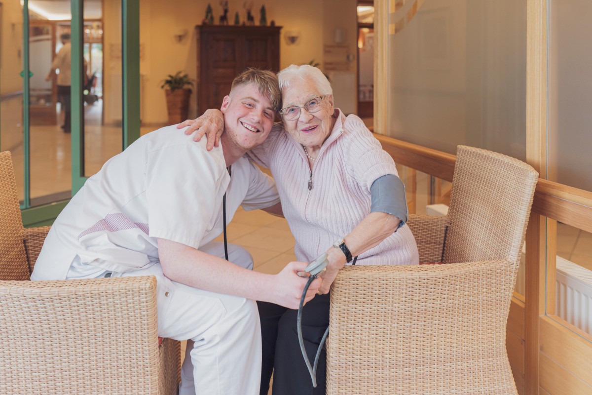 Friendly young nurse in white uniform hugging smiling elderly woman in nursing home, both sitting in rattan chairs, woman wearing blood pressure cuff, radiating warm and trusting atmosphere.