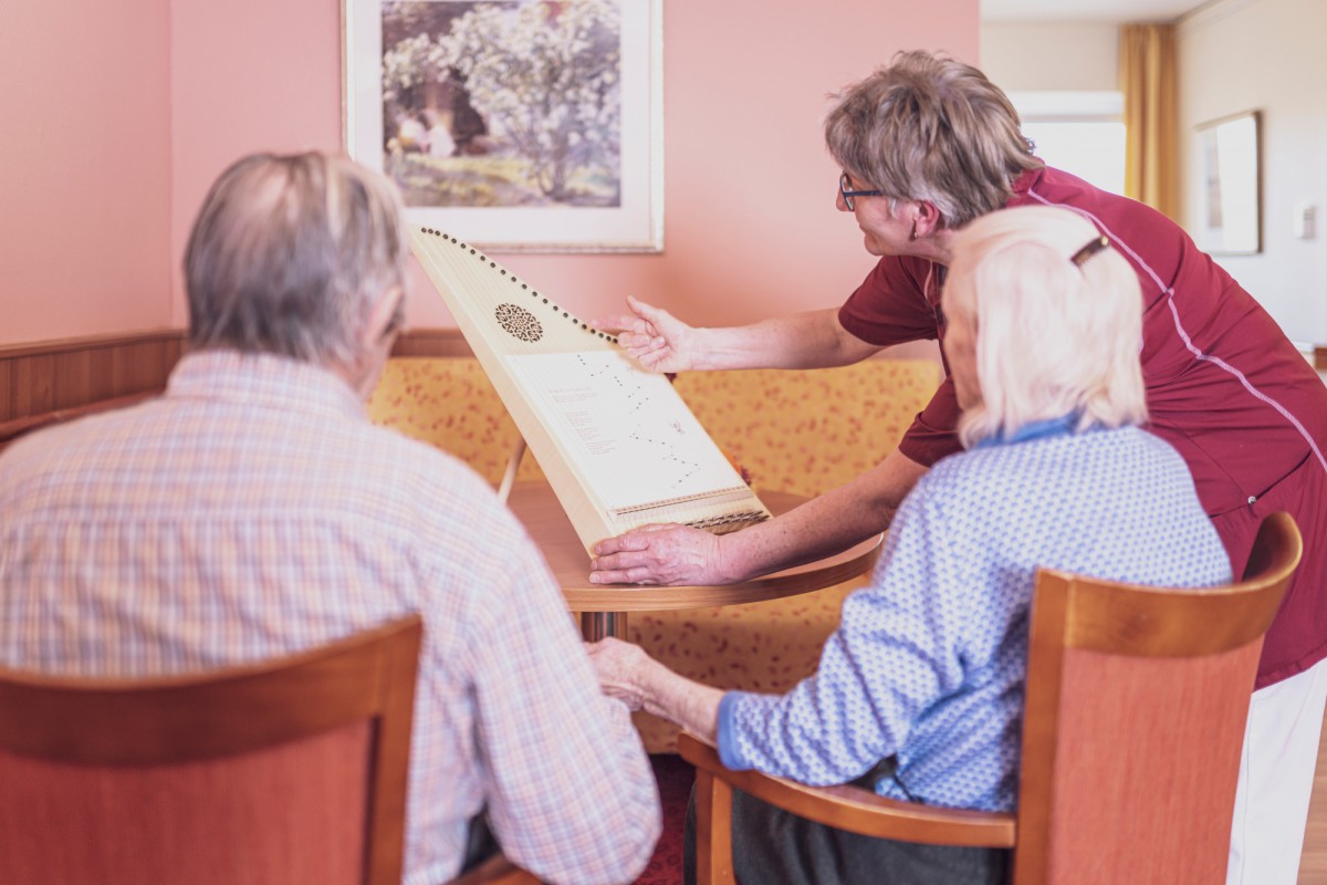 Professional in red clothing explains something on a board with dot patterns to two elderly people while sitting around a table in a cozy setting.
