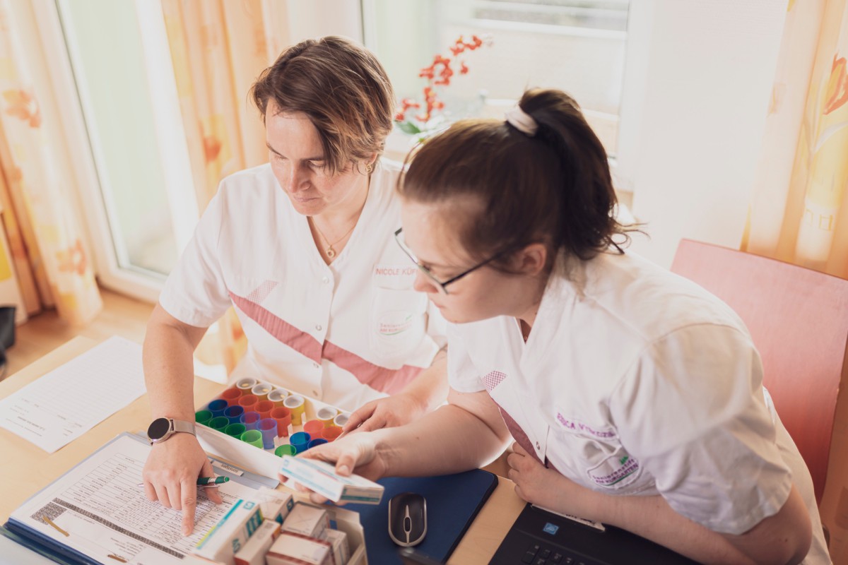 Two female employees in white professional clothing working concentratedly with a rack of colorful tubes and documents at a table.