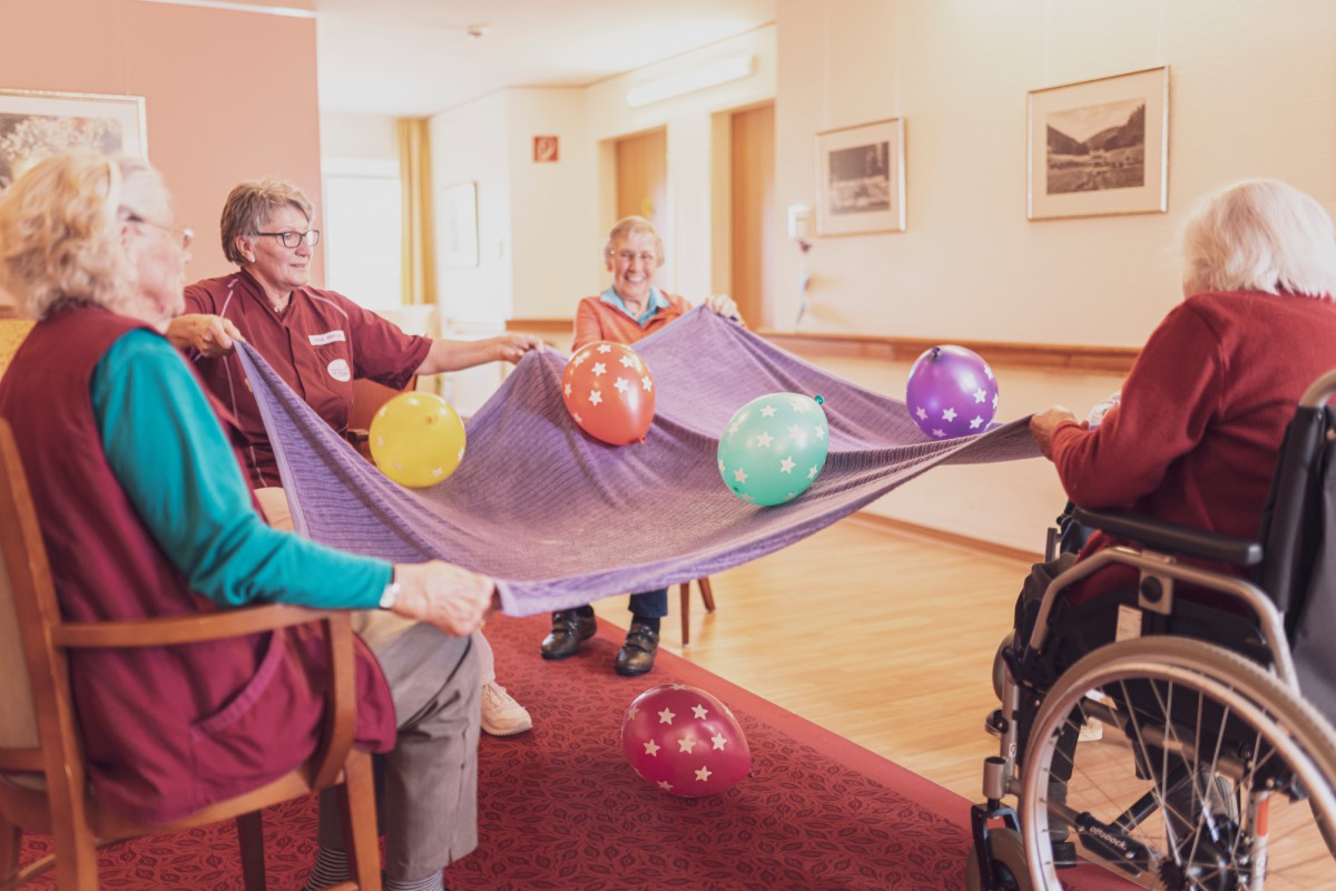 Elderly individuals in a care facility engage in a group activity, holding a stretched purple cloth with colorful balloons balanced on top, seated in chairs and a wheelchair in a bright, spacious room.