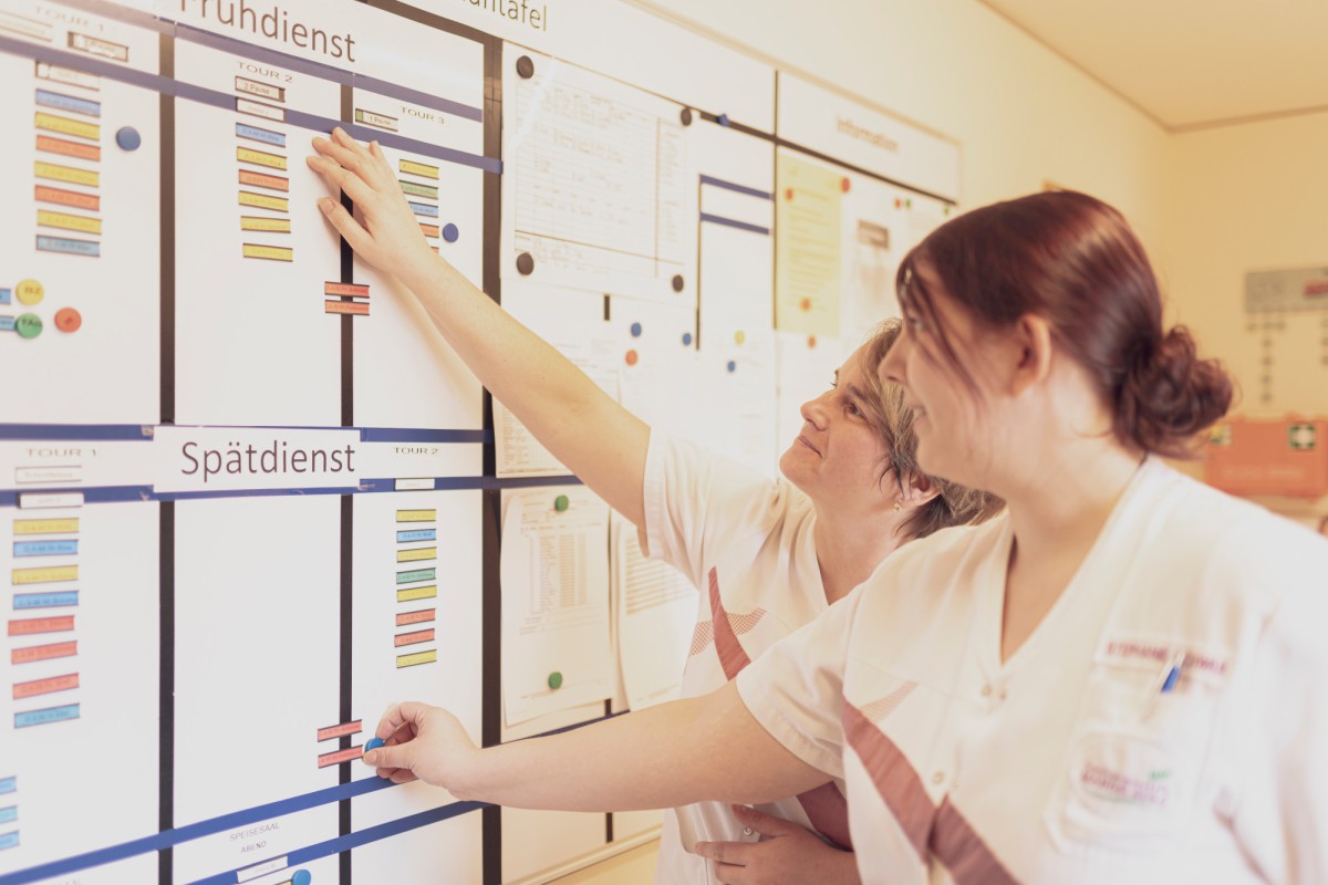 Two people in white professional attire stand before a shift schedule board, arranging colored shifts and time blocks for different services like "morning shift" and "late shift".
