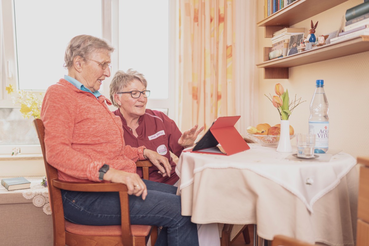 Two people sitting at a table with a red tablet, surrounded by a cozy living room atmosphere with books, curtains, and flowers.