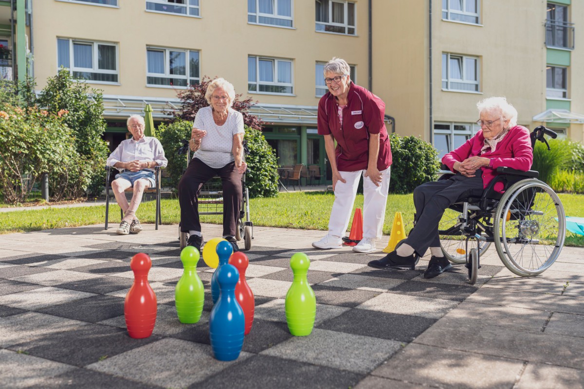 Three seniors playing bowling outdoors on a checkered surface, with a staff member in red and white assisting them.