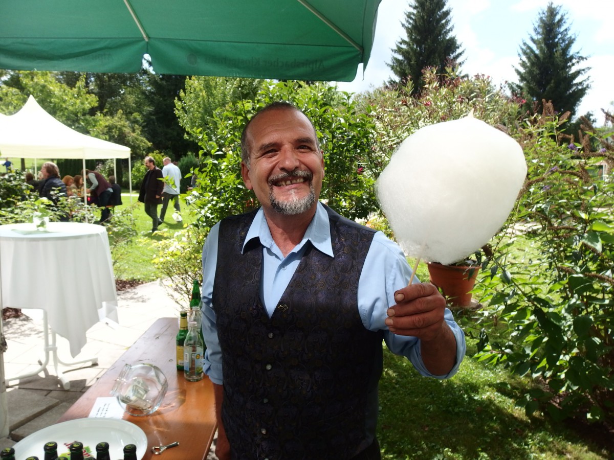 A friendly man in a vest and shirt smiles widely while holding cotton candy towards the camera.