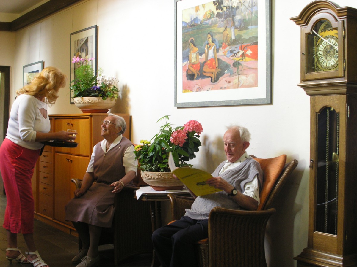 Two elderly people in a homey living room with wooden furniture, flowers, and a wall clock. A woman hands a glass of water.