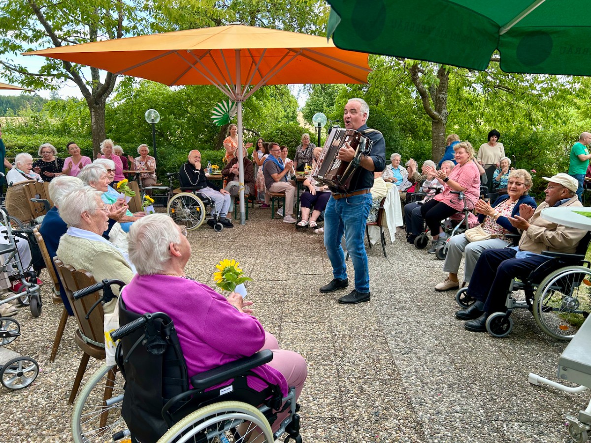Elderly men and women sitting under orange and green umbrellas while a man plays accordion, creating a relaxed outdoor atmosphere.