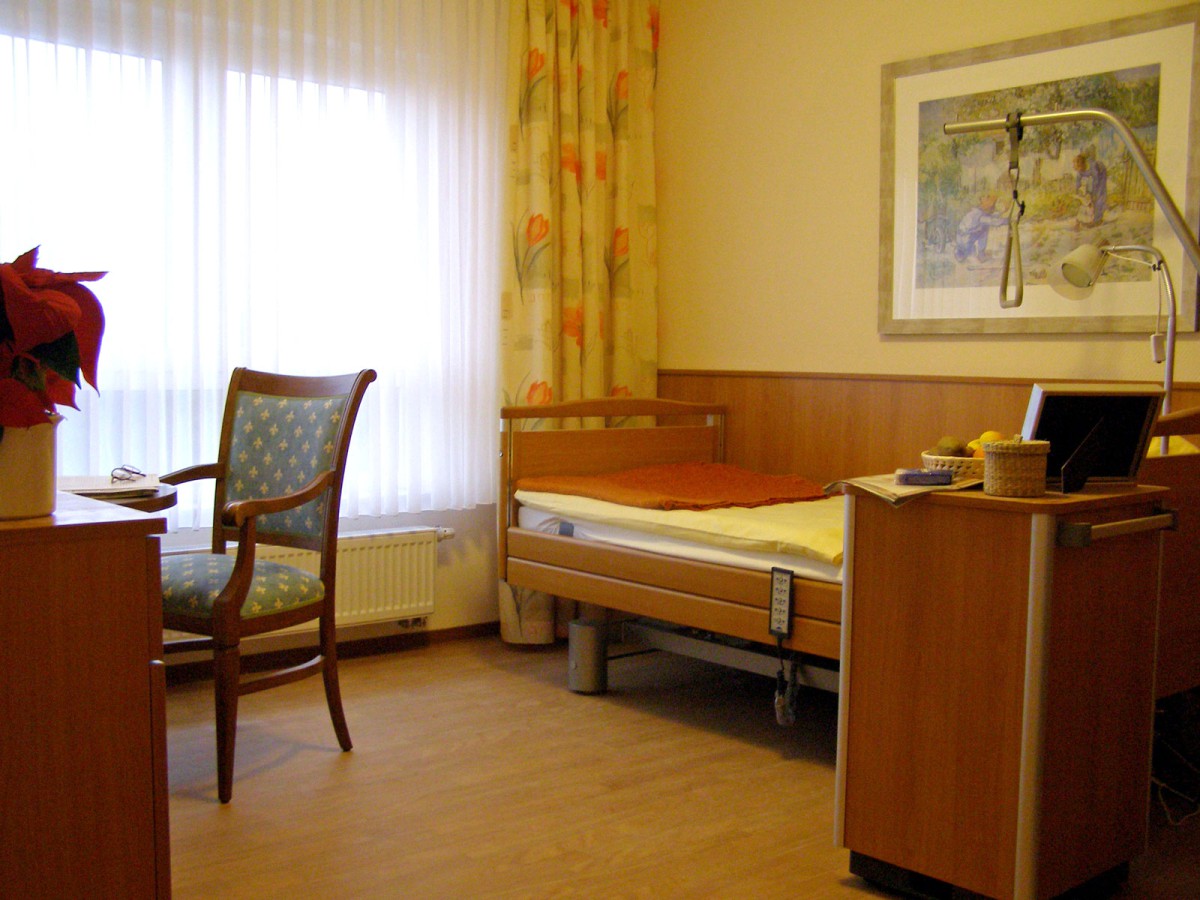 Room with a single bed, adjustable nightstand, chair, and a window with curtains. An art print hangs on the wall.