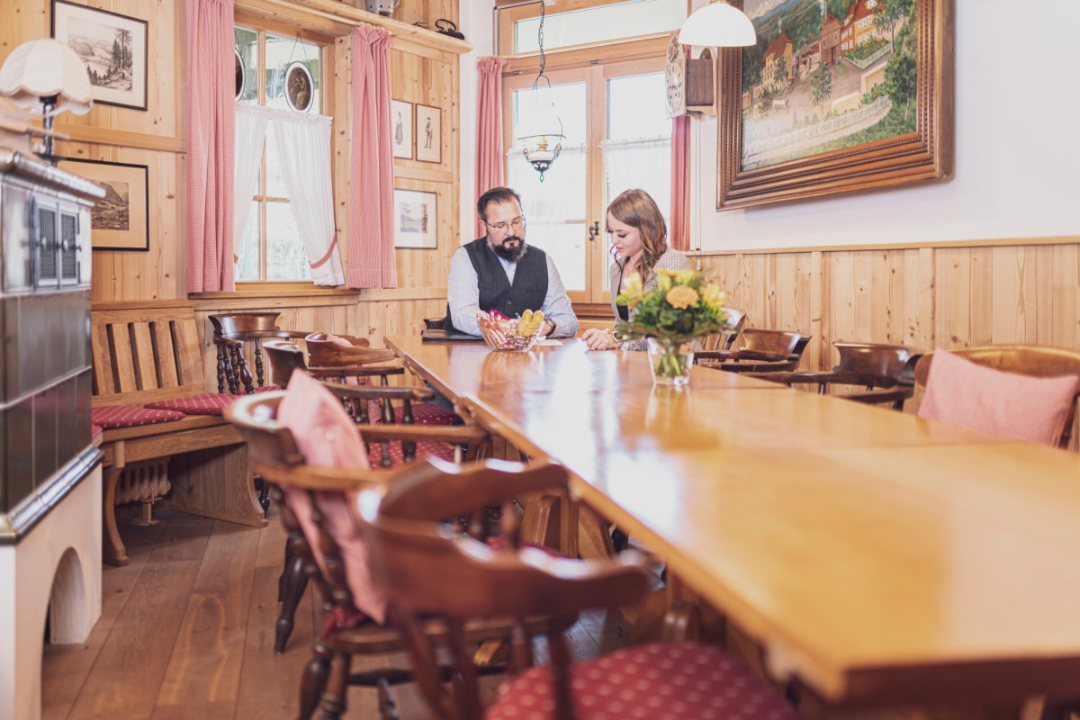 An elegant dining room with wood paneling, where two people sit at a long wooden table, surrounded by rustic wooden chairs and paintings on the walls.