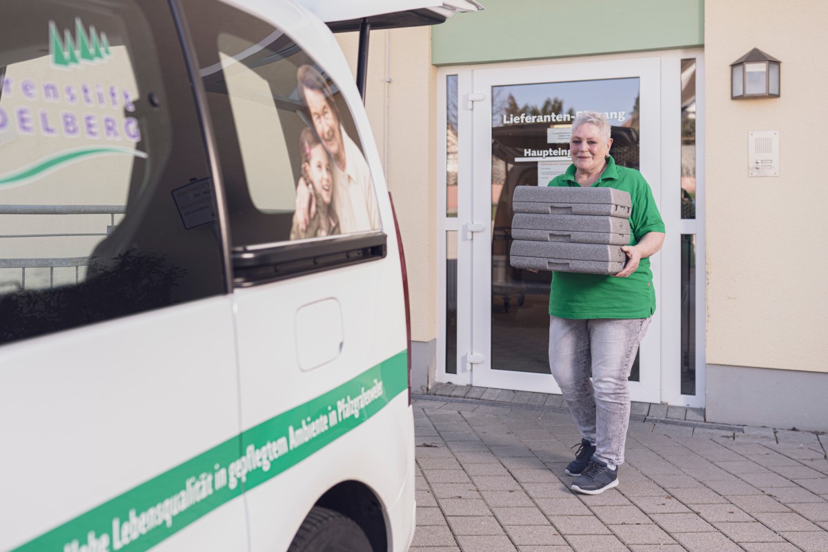 A person in a green t-shirt carrying multiple stacked packages in front of a white delivery van with green lettering, near a building entrance with a glass door.