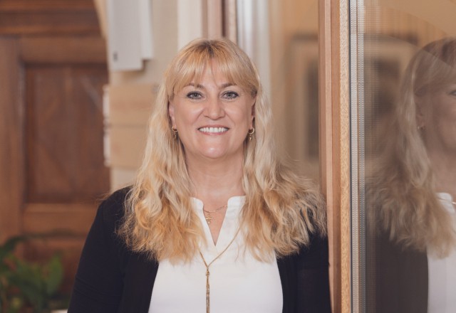 Smiling middle-aged blonde woman with bangs and long hair, wearing a white blouse and black cardigan, standing indoors with wooden door in background.