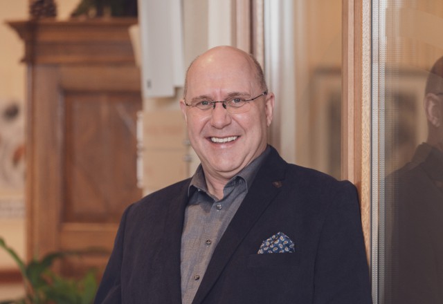 Smiling bald man with glasses wearing a black blazer and gray shirt, standing indoors with a friendly, positive demeanor.