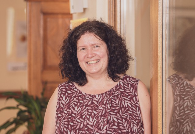 A smiling middle-aged woman with shoulder-length curly black hair wearing a sleeveless top with a leaf pattern in red and white tones.