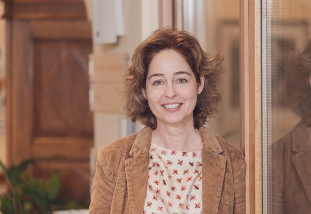 Smiling woman with wavy brown hair, wearing a cream floral top and brown corduroy vest.