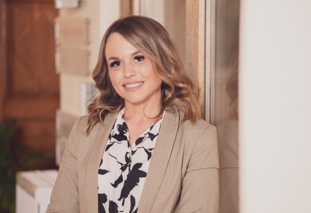 A smiling woman with wavy, shoulder-length hair in a beige blazer over a black and white patterned top, standing in front of a wooden door and smiling warmly at the camera.