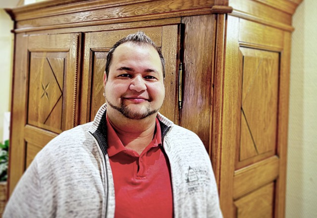 Man in red polo shirt and gray marled cardigan smiles friendly at camera, with large wooden cabinet in background.